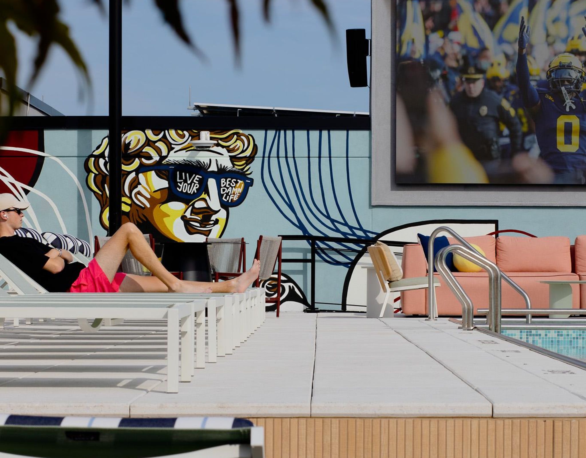 Verve Ann Arbor man relaxing by a pool with wall art and a large screen showing a football game in the background.