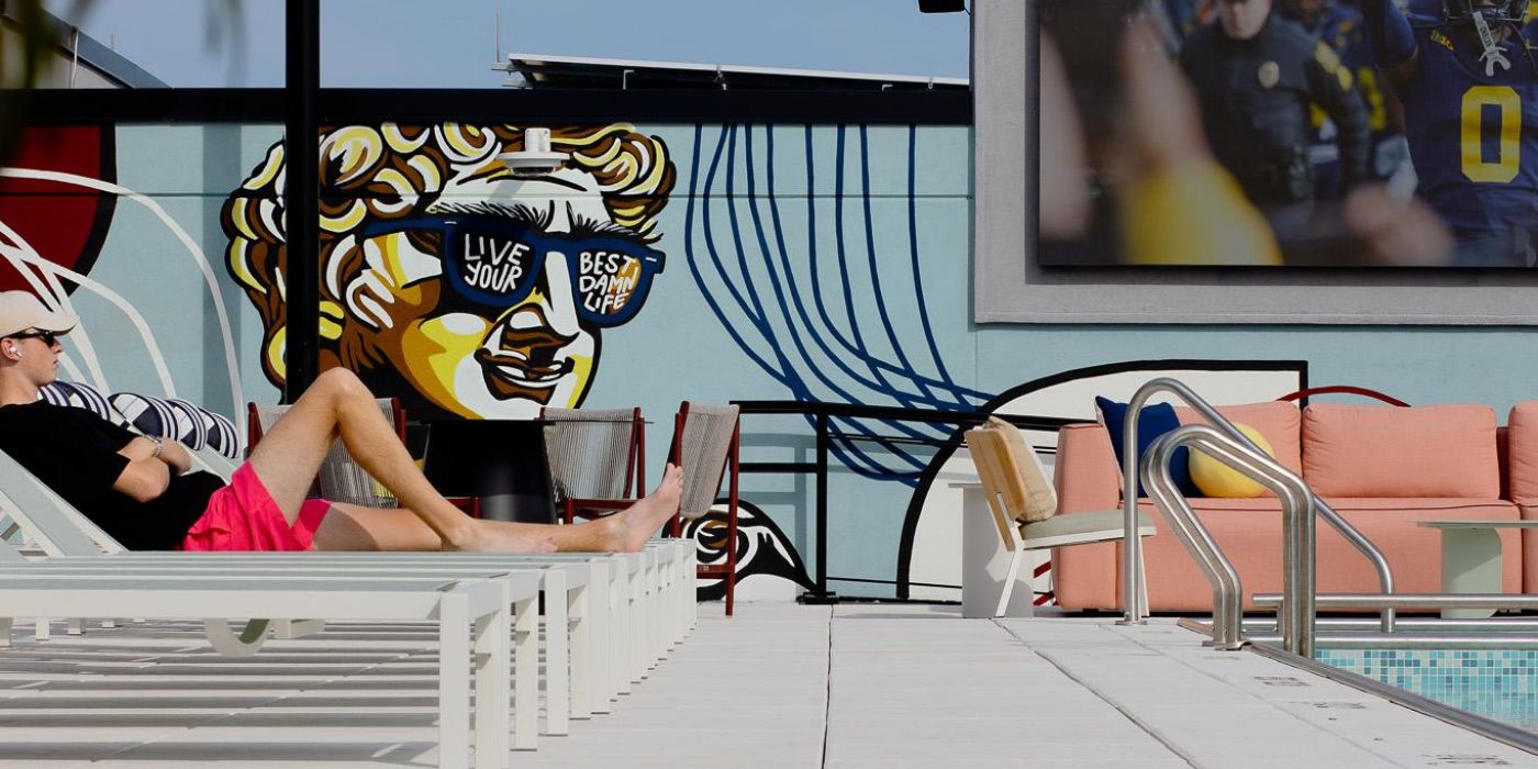 Verve Ann Arbor man relaxing by a pool with wall art and a large screen showing a football game in the background.